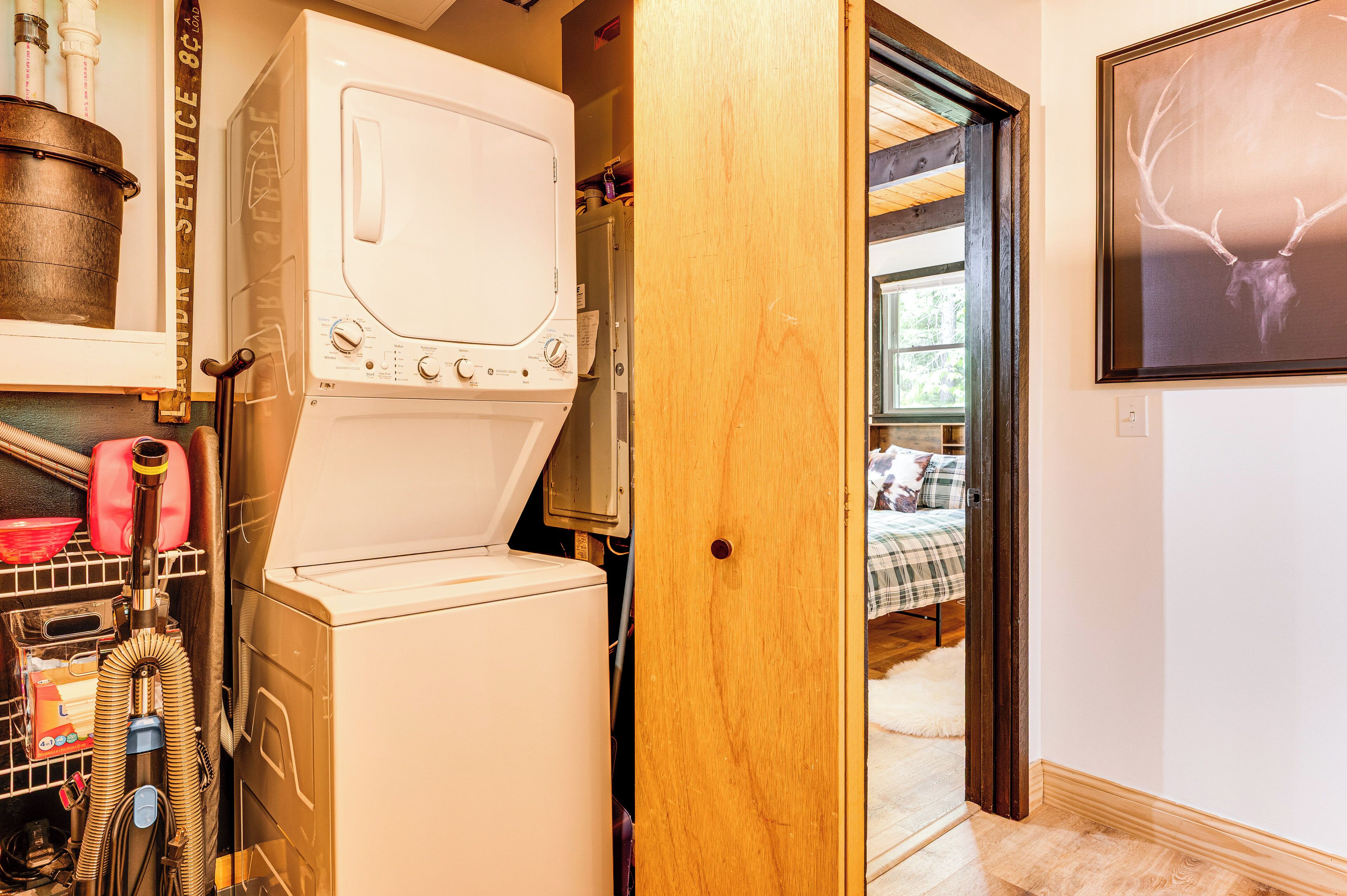 A laundry room with a washing machine and dryer, a vacuum cleaner, and a wooden door leading to a bedroom with a bed and a framed deer head picture on the wall.
