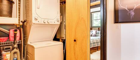 A laundry room with a washing machine and dryer, a vacuum cleaner, and a wooden door leading to a bedroom with a bed and a framed deer head picture on the wall.