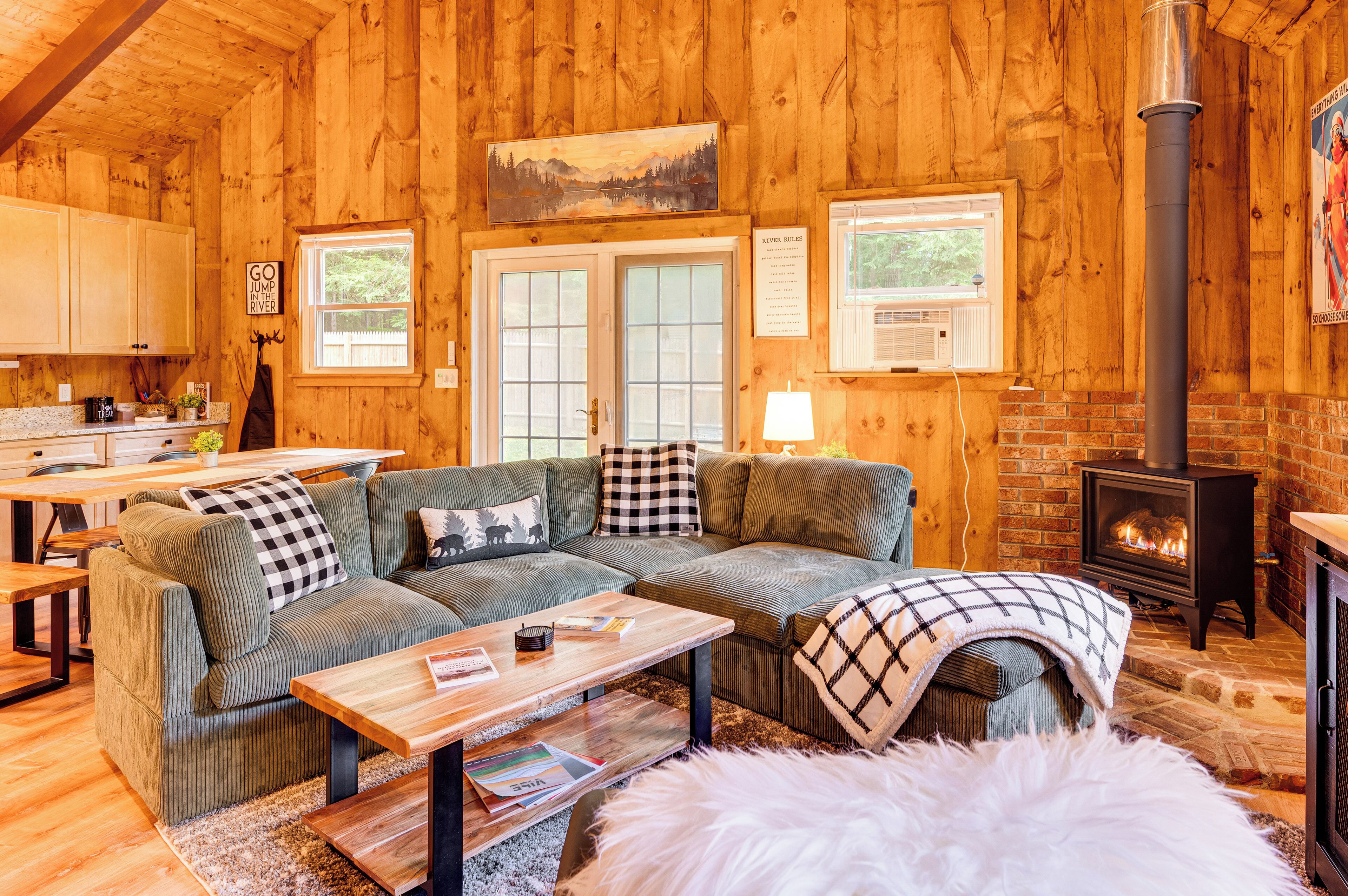 A cozy cabin interior with a sectional sofa, a fireplace, wooden walls, and a kitchen area.