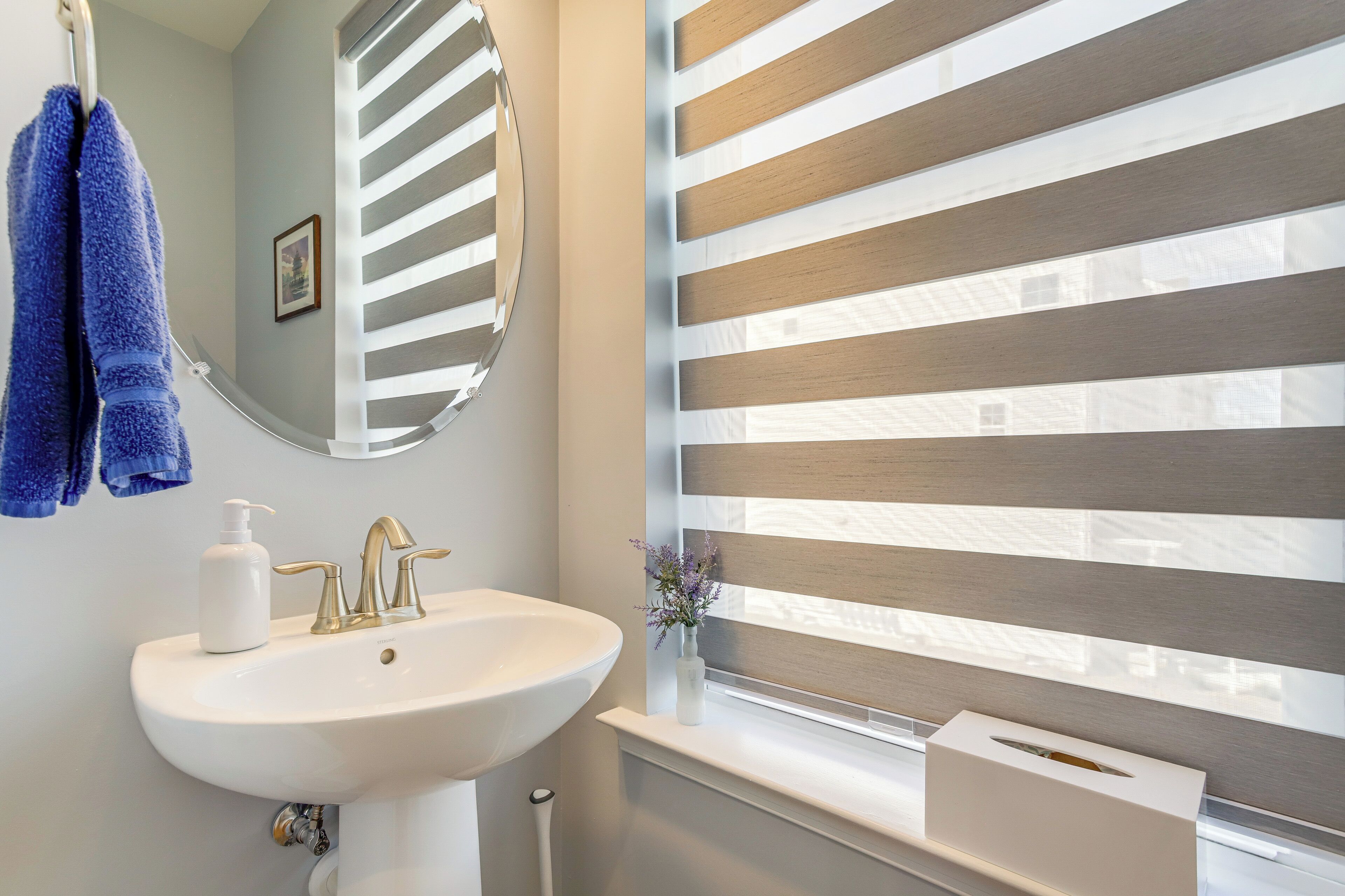 Une salle de bain avec un lavabo blanc, des robinets dorés, un miroir rond et une fenêtre avec des stores horizontaux.