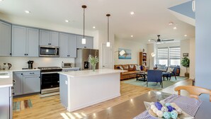 A modern kitchen with grey cabinets, a central island, and a dining area with a table and chairs. - Walk to Dtwn: Waterfront Eastern Shore Townhome! (Cambridge)