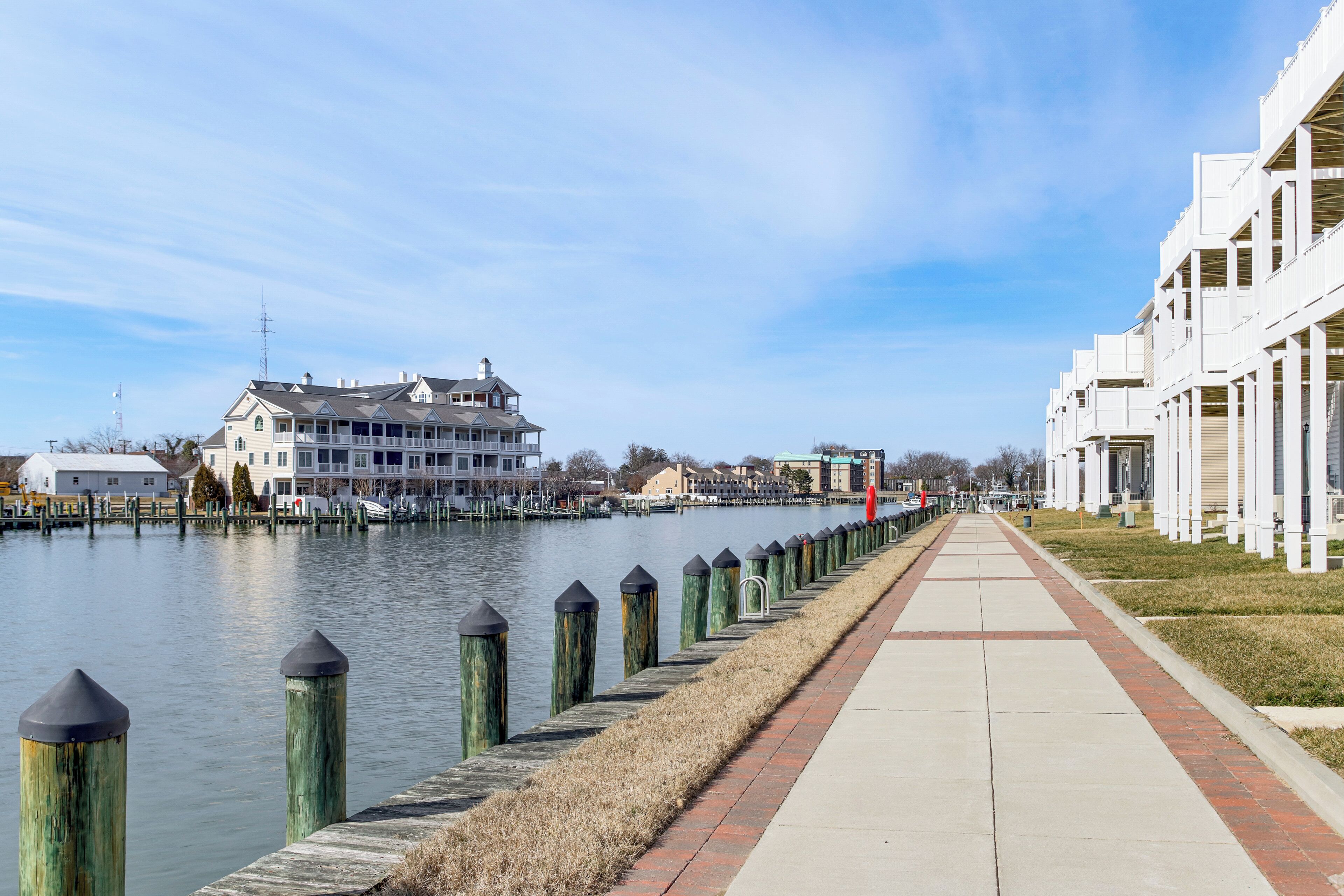 A waterfront promenade with a row of buildings, a clear sky, and a calm water body.