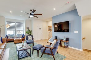 A modern living room with a ceiling fan, a flat-screen TV mounted on the wall, a sofa set, and a coffee table. - Walk to Dtwn: Waterfront Eastern Shore Townhome! (Cambridge)