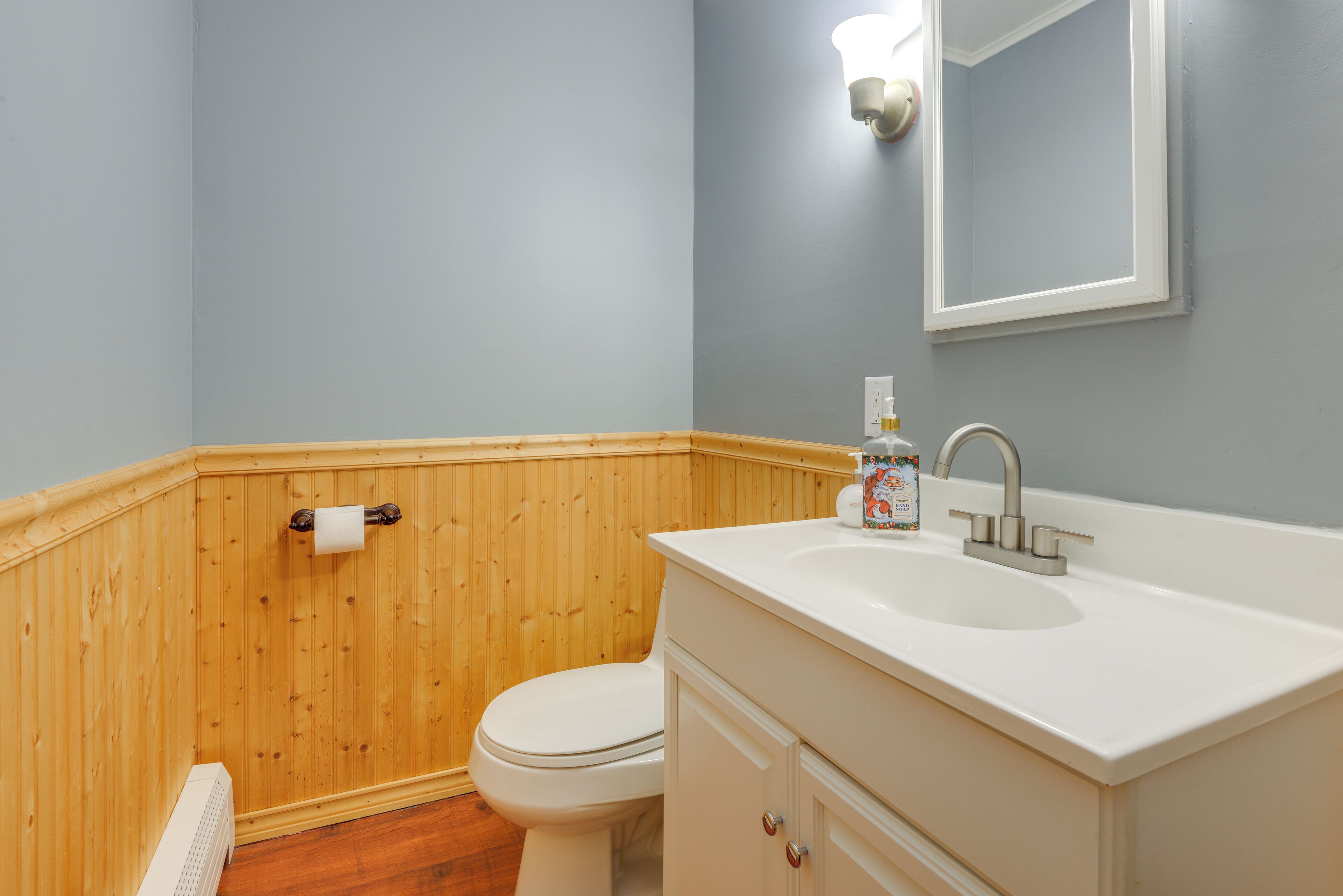 Une salle de bain avec un mur recouvert de panneaux en bois, des toilettes blanches, un lavabo blanc avec un miroir au-dessus, et un flacon de savon liquide sur le lavabo.