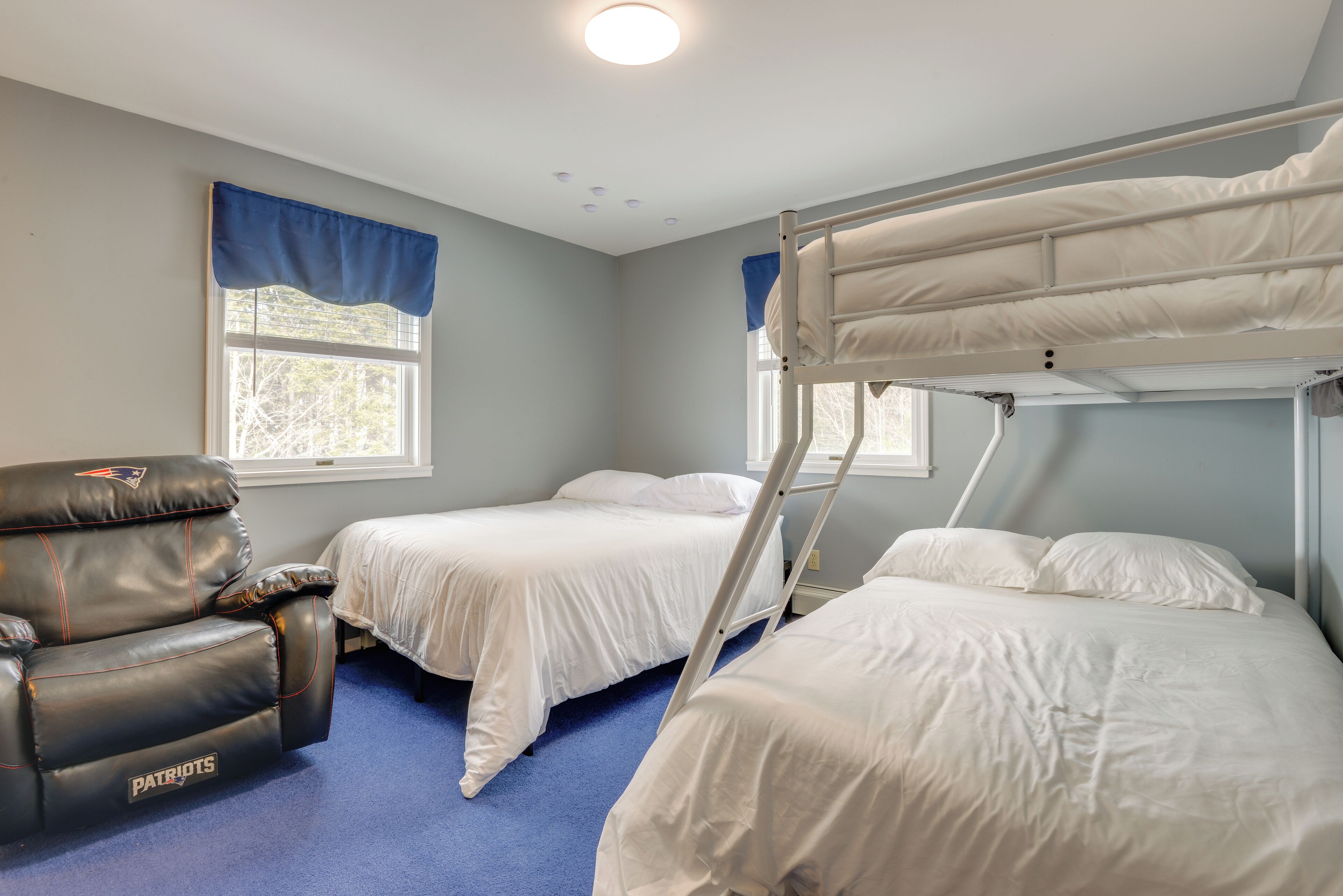 A room with bunk beds, a single bed, a leather chair with a logo, and a window with blue curtains.