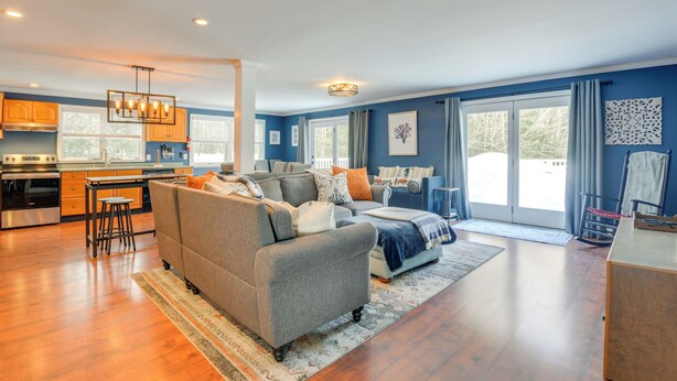 A spacious living room with a grey sofa, a dining area with a table and chairs, and a kitchen with wooden cabinets and a stainless steel range hood.