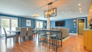 A modern living room with a dining area, a large TV mounted on the wall, and a kitchen area with wooden cabinets.