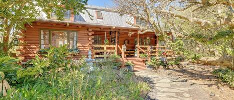 A log cabin with a porch, surrounded by lush greenery and a stone pathway.