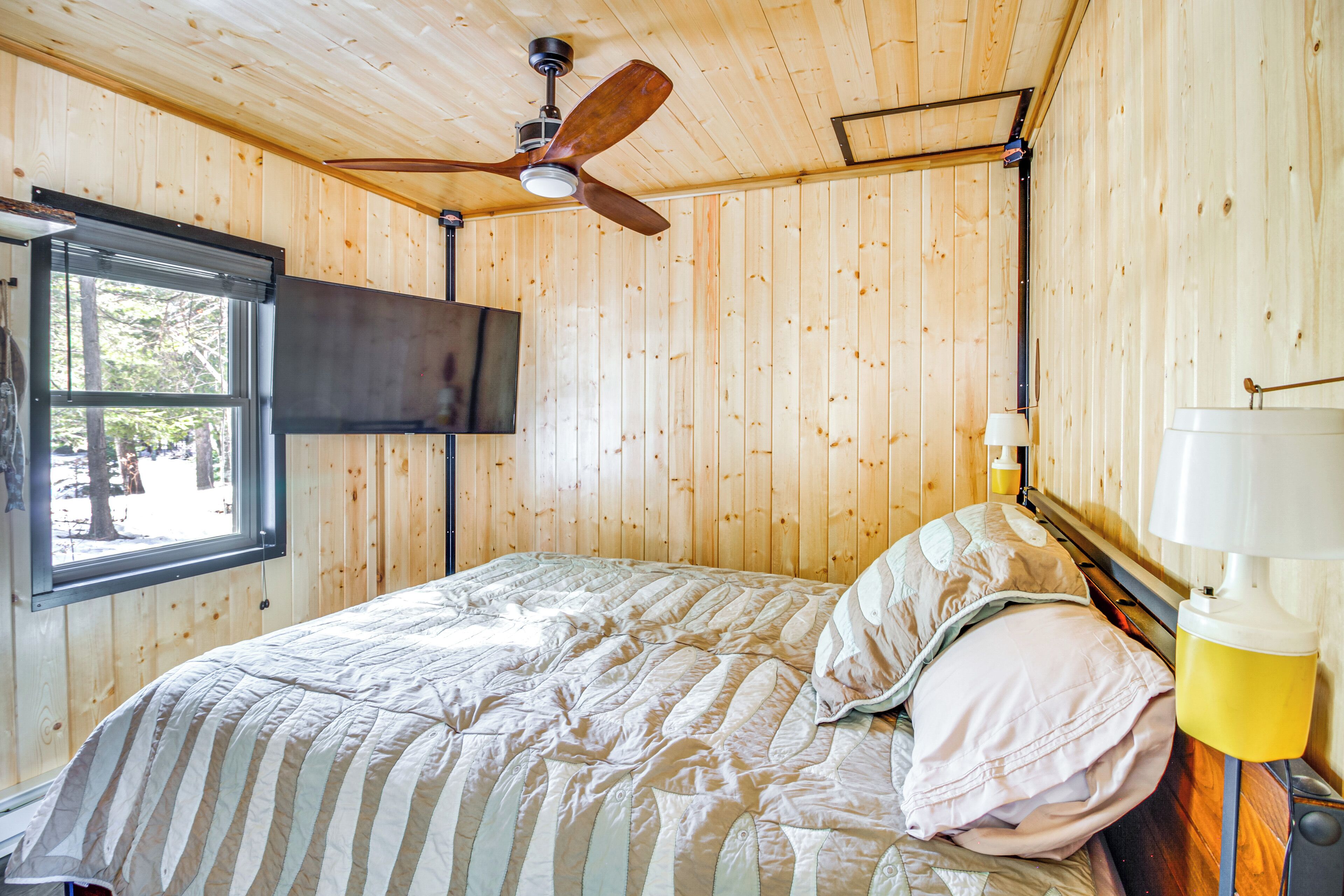A wooden-paneled room with a bed, a ceiling fan, a window, and a TV mounted on the wall.