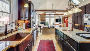A spacious kitchen with dark wood cabinets, a large island with a sink, and a red rug. There is a stone fireplace and a large window providing natural light. - Large Deck: Luxe 6-acre Retreat in Dahlonega! (Dahlonega)