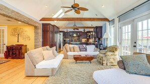 A spacious living area with a high ceiling, wooden beams, and a large ceiling fan. There is a cozy seating area with a sofa and armchairs, and a kitchen visible in the background. - Large Deck: Luxe 6-acre Retreat in Dahlonega! (Dahlonega)
