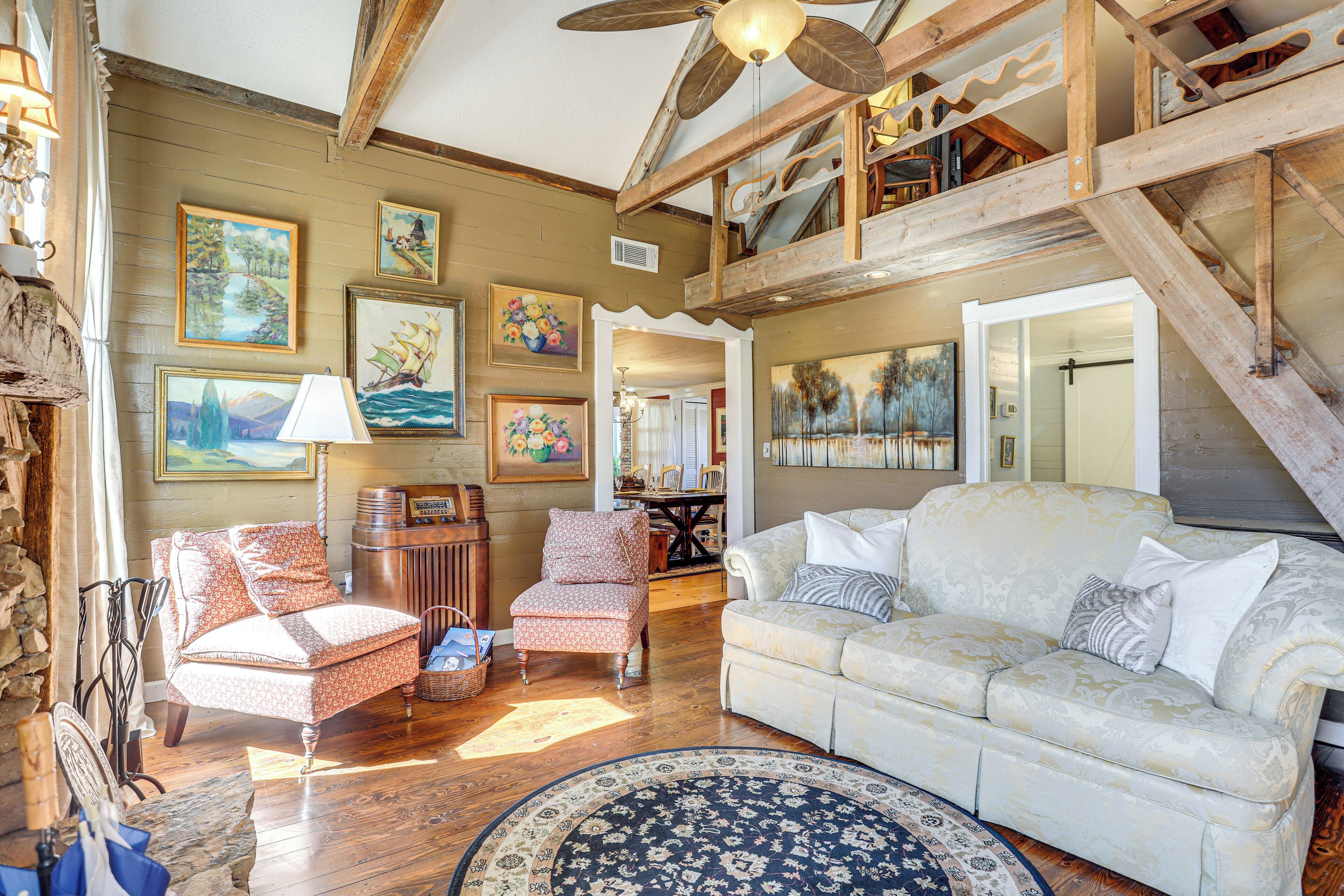 A spacious living room with a high ceiling, wooden beams, a large sofa, a patterned rug, and various framed artworks on the walls.