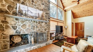 A stone fireplace with a TV, a wooden ceiling fan, and a cozy seating area with a rug and a couch. - Golf, Fish & Ski: Retreat w/ Game Room in Thompson (Thompson)