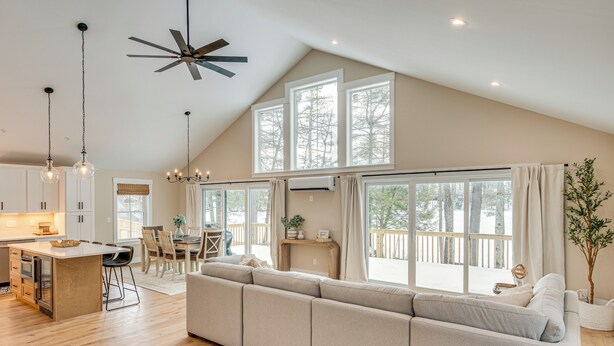 A spacious living area with a large sofa, wooden flooring, a kitchen island, and a dining area with natural light from large windows. - Dock & Game Room: New Waterfront Home in Lyman! (Alfred)