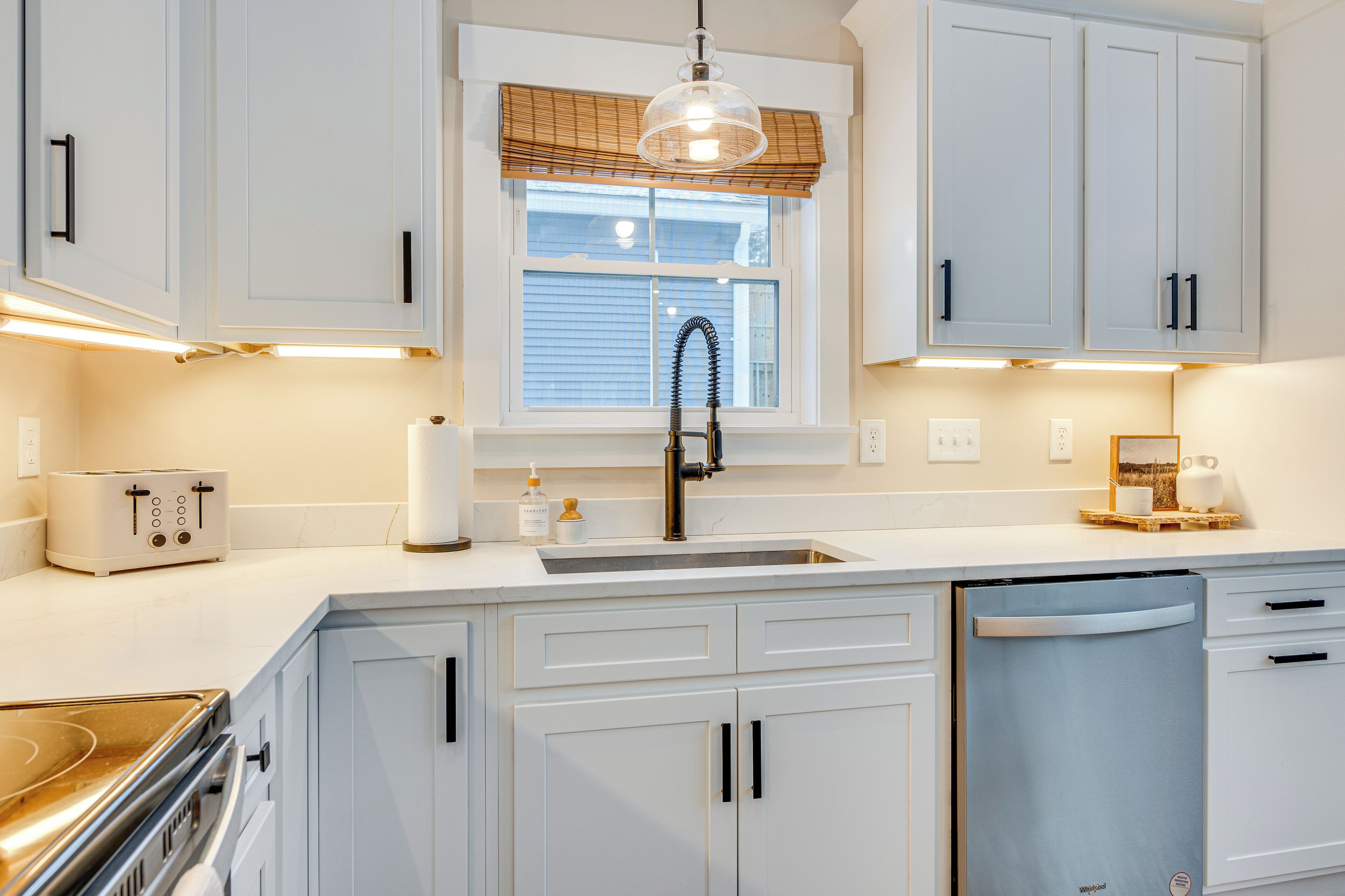 A modern kitchen with white cabinets, a stainless steel range hood, a built-in dishwasher, and a window with blinds.