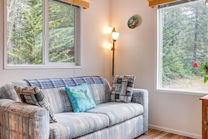 A room with a sofa, two windows, a lamp, and a view of a forest. - Remote Workers Welcome: Tiny Home in Port Angeles! (Port Angeles)