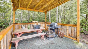 Eine überdachte Holzterrasse mit Picknicktischen und Grill, umgeben von Wald.