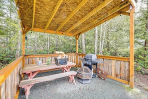 A covered wooden deck with picnic tables and a grill, surrounded by forest. - Remote Workers Welcome: Tiny Home in Port Angeles! (Port Angeles)