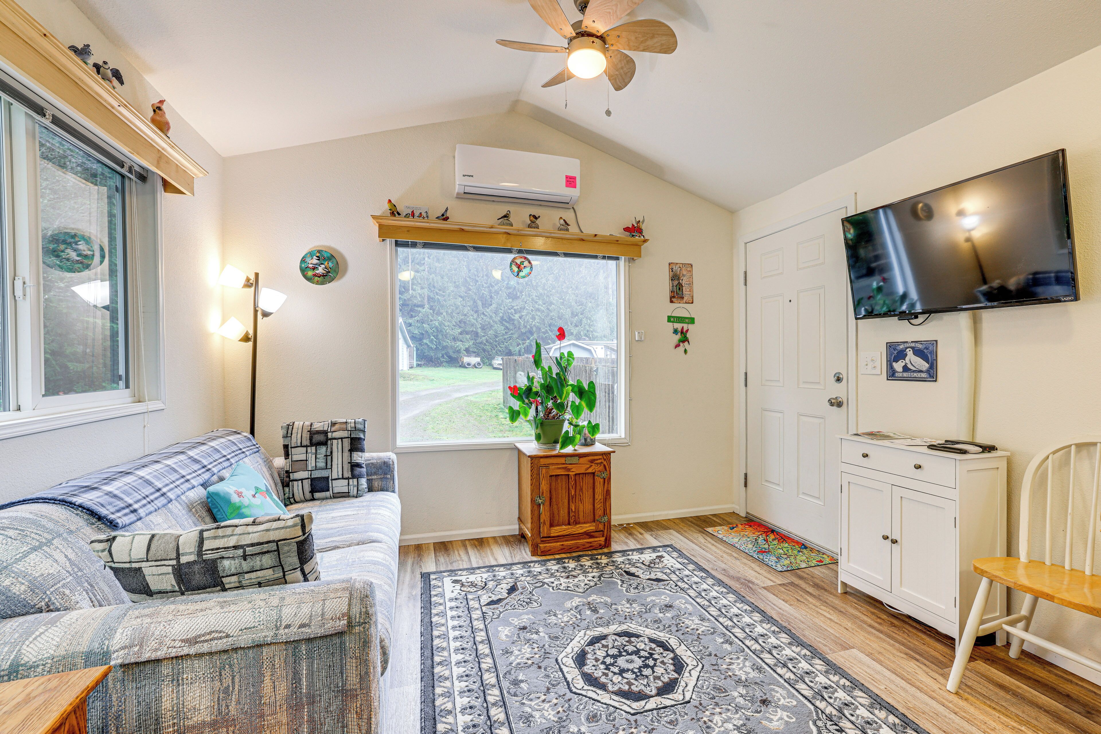 A cozy living room with a sofa, a small wooden cabinet, a television, and a window with a view of a garden.