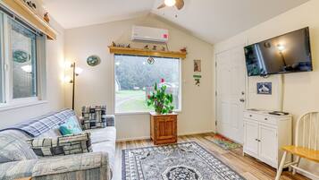 A cozy living room with a sofa, a small wooden cabinet, a television, and a window with a view of a garden.