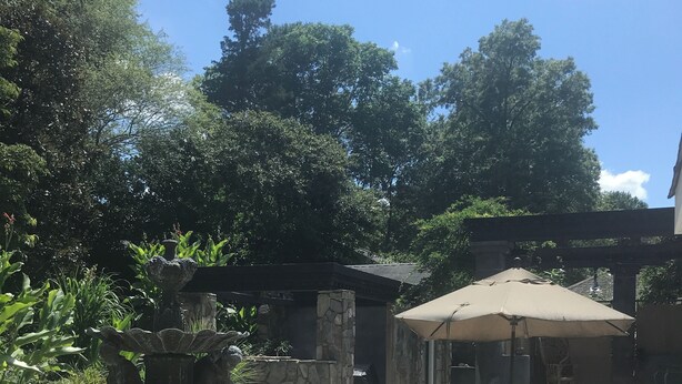 Un espace extérieur aménagé en terrasse, avec une fontaine en pierre, des sièges à l’abri d’un auvent et une végétation luxuriante tout autour.