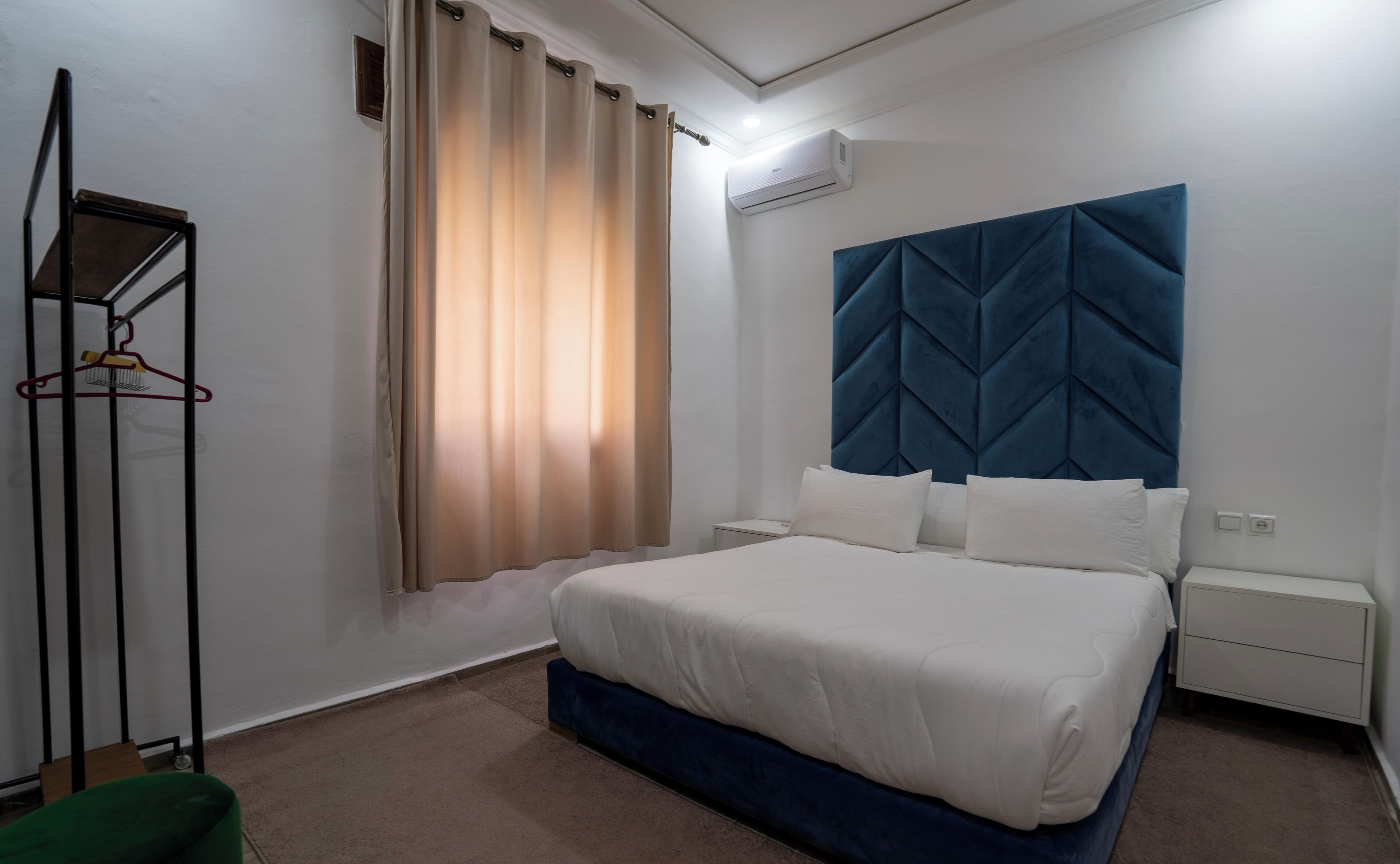 Un dormitorio con una cama, cabecera, mesita de noche, perchero y ventana con cortinas.