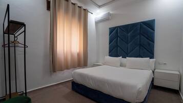 Un dormitorio con una cama, cabecera, mesita de noche, perchero y ventana con cortinas.