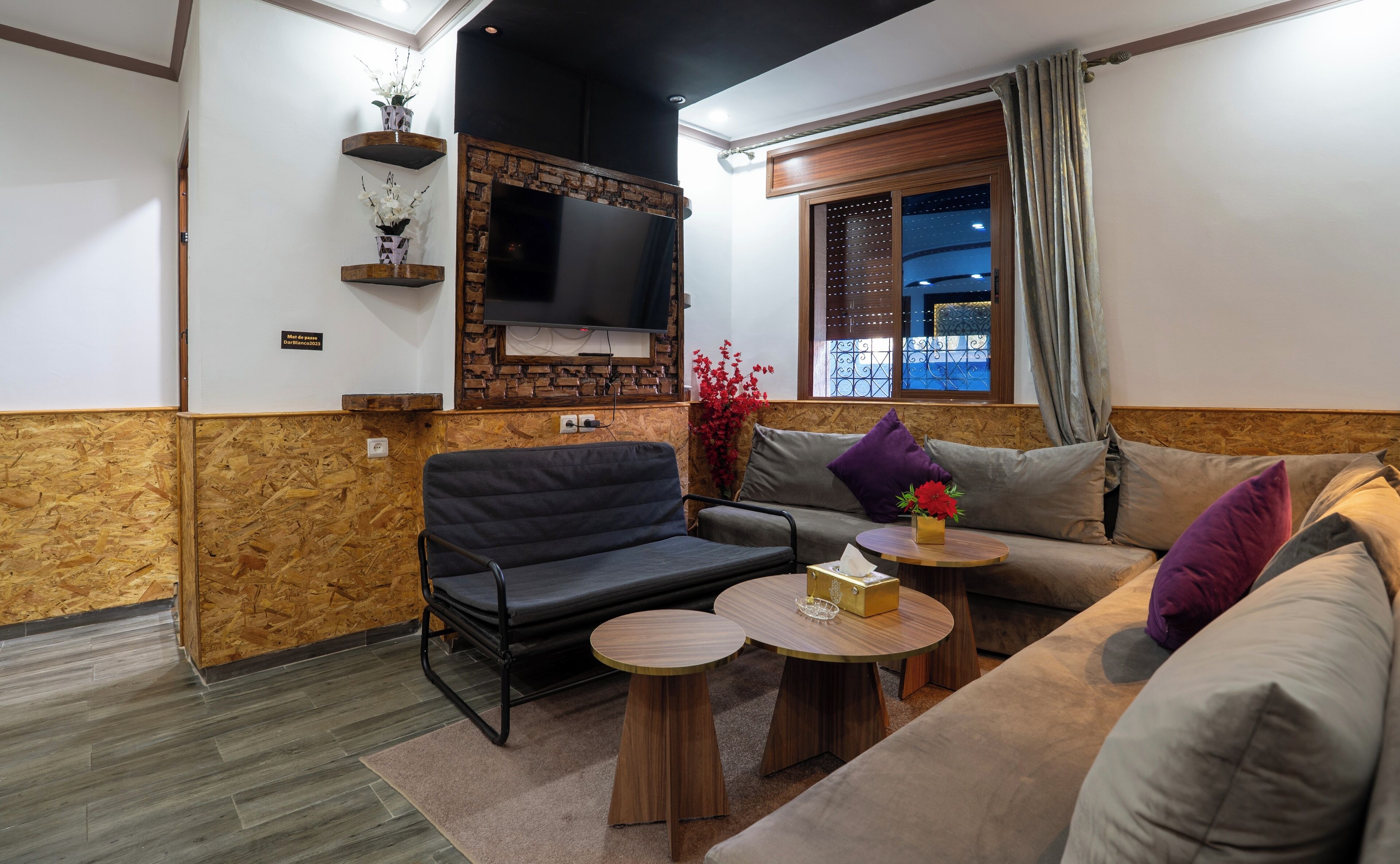 A living room with a sofa, two armchairs, a coffee table, a TV mounted on a brick wall, and a window with curtains.