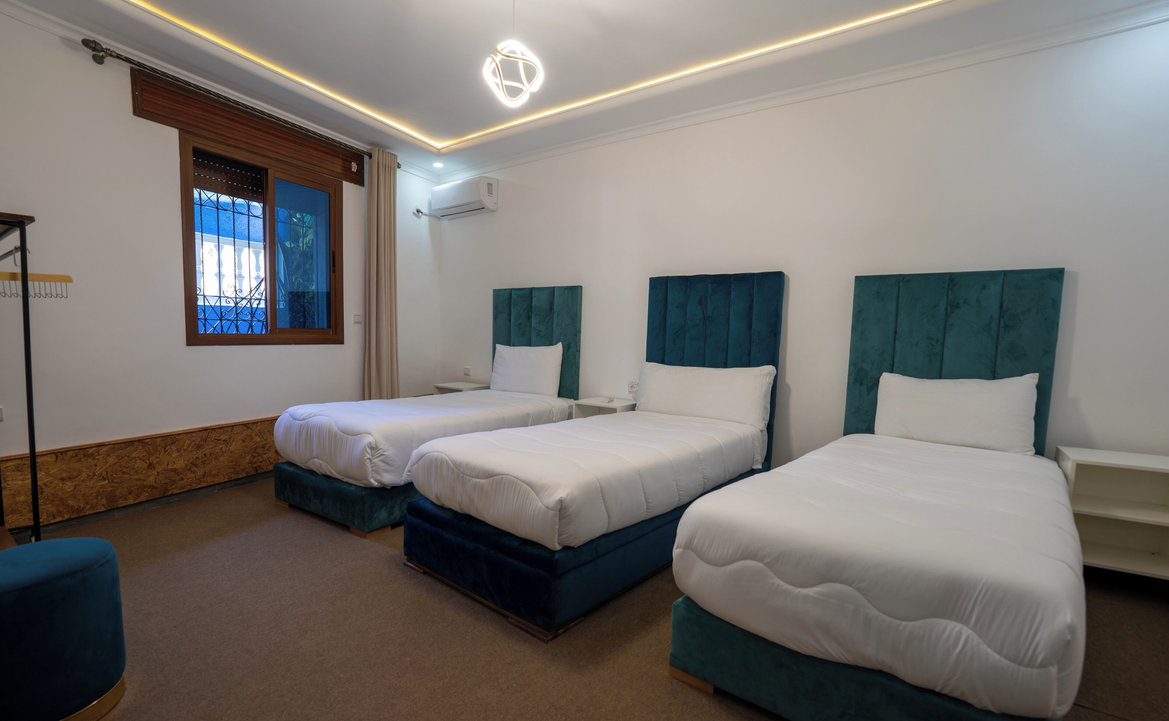 A hotel room with two beds, each with a teal headboard and white bedding. There is a window with a view of greenery outside, and an air conditioning unit mounted on the wall.