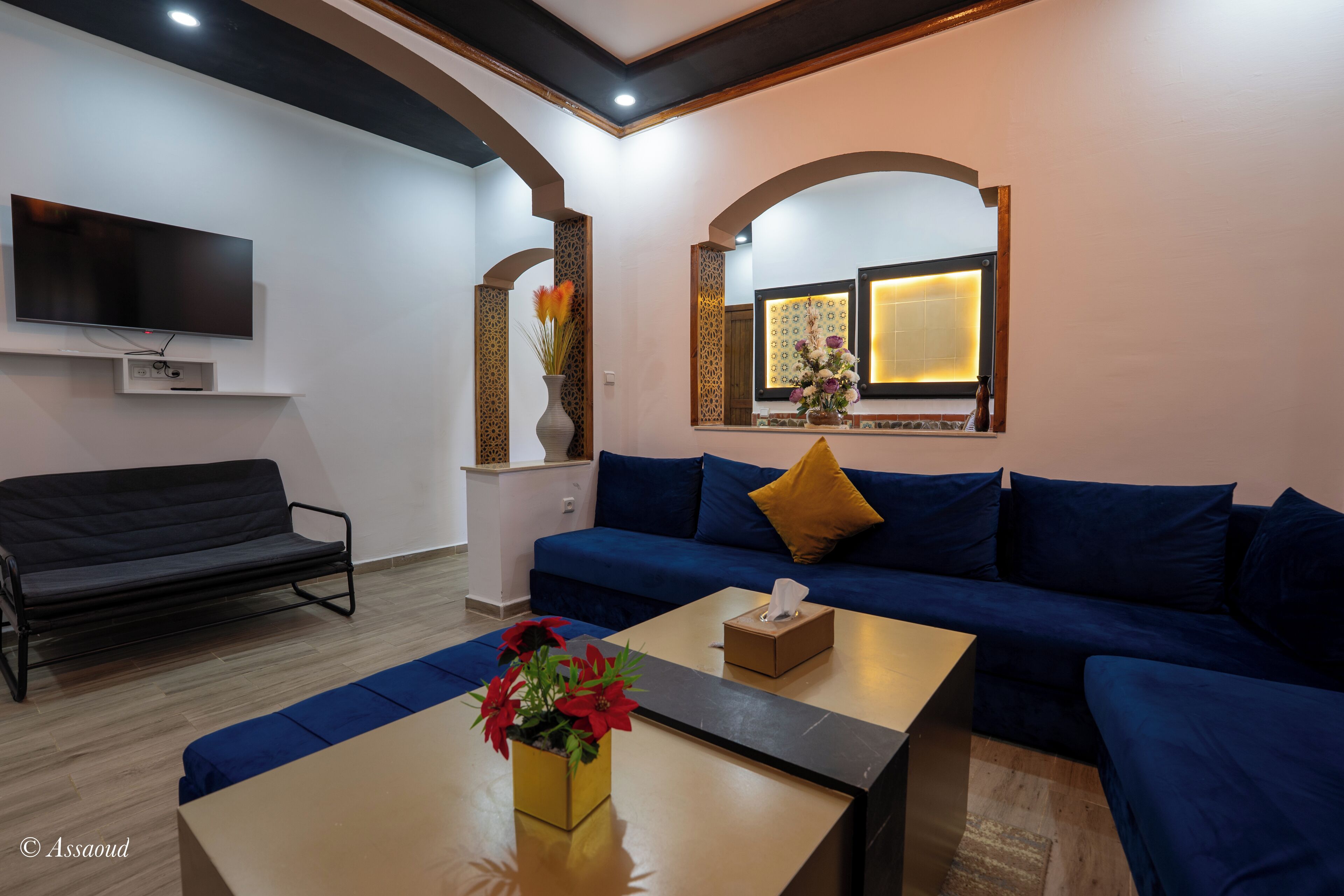 A living room with a blue sofa, a coffee table with a vase of flowers, a flat-screen TV mounted on the wall, and a window with decorative lighting.