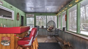 A bait shop with a wooden counter, red chairs, a hanging chair, and a view of trees through large windows.