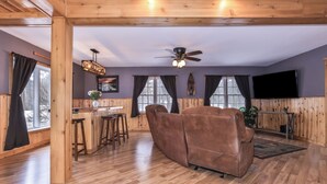 A cozy room with a wooden ceiling, a ceiling fan, a bar area with stools, a brown leather sofa, and a TV mounted on the wall.