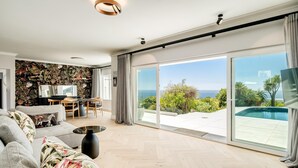 Een moderne woonkamer met een grote bank, een salontafel en een televisie. Er is een eethoek met een tafel en stoelen. De kamer heeft grote schuifdeuren die leiden naar een balkon met een zwembad.