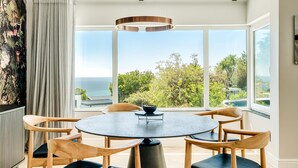 Een eetruimte met een ronde tafel en zes houten stoelen, grote ramen met uitzicht op de oceaan, en een moderne hanglamp.