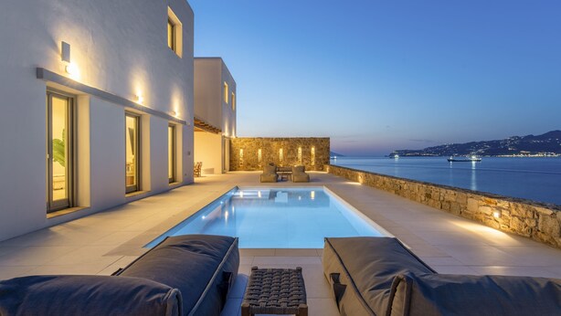 Ein moderner Poolbereich mit freiem Blick auf das Meer, einer Steinmauer und einem Gebäude mit großen Fenstern.