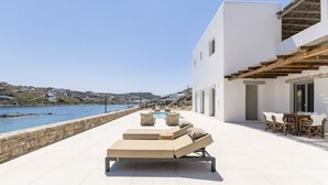 Eine moderne Lounge-Bereich im Freien mit Pool, beige Liegestühlen und einem Essbereich mit Blick auf eine Küstenlandschaft.