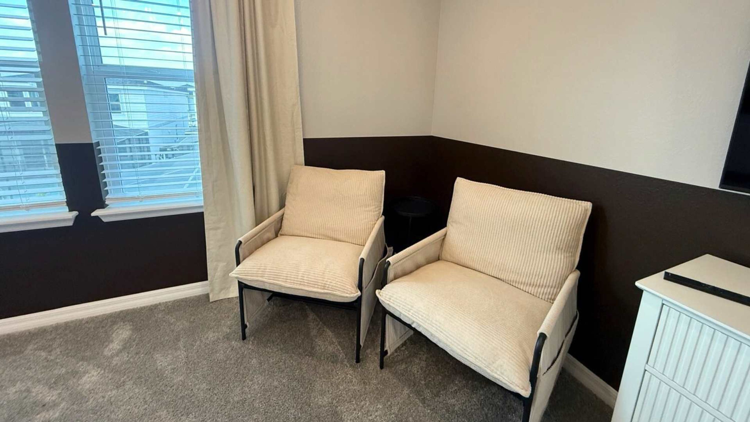 Ett hörnrum med två beige fåtöljer, ett litet bord och ett element.