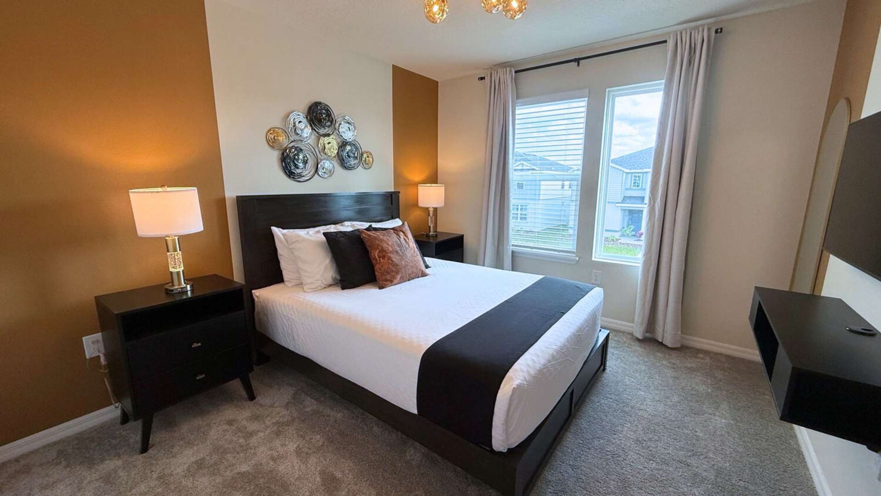 A neatly arranged bedroom with a large bed, two bedside tables with lamps, a wall clock, and a window with blinds.
