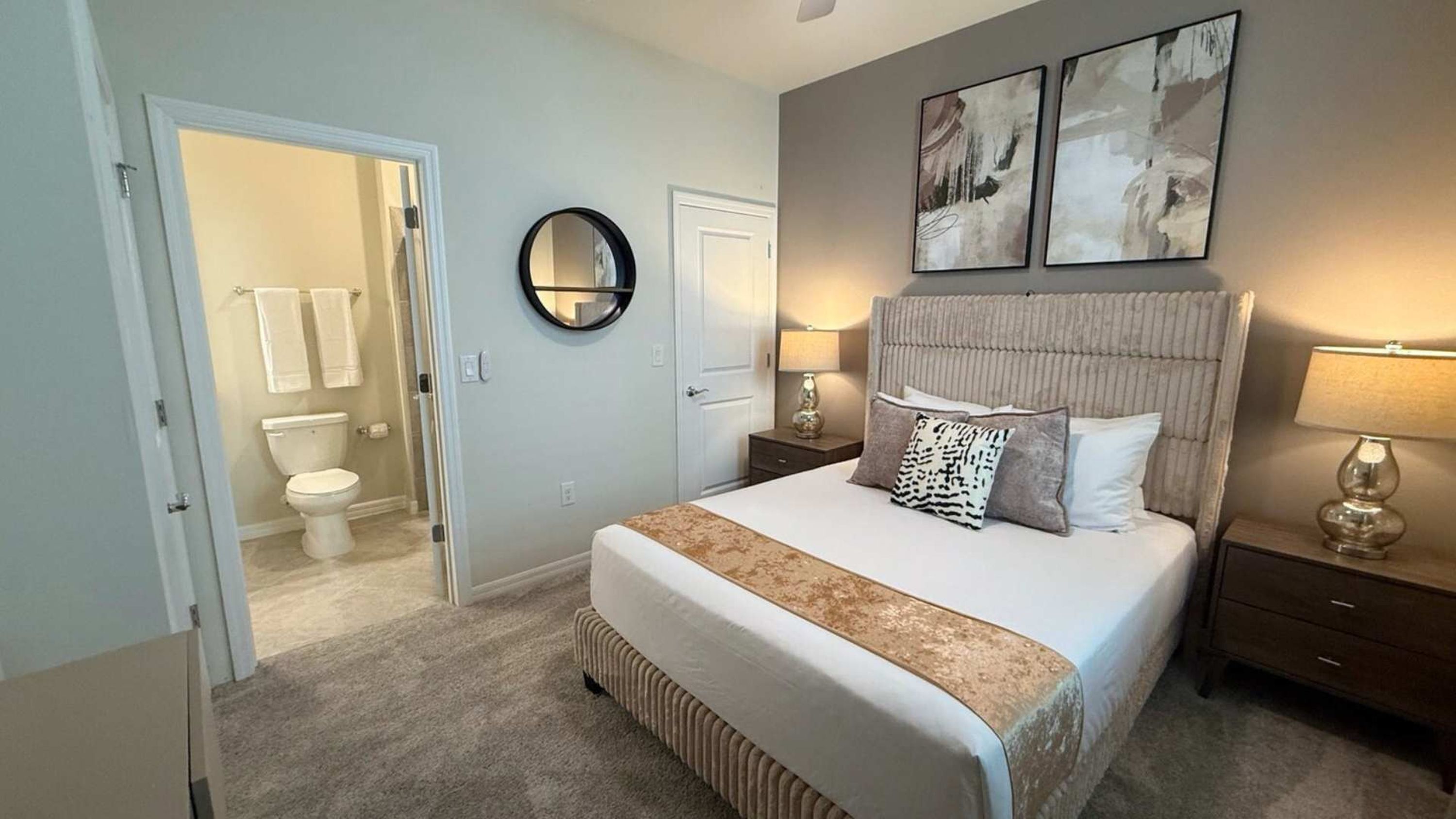 A bedroom with a large bed, a nightstand with a lamp, a round mirror, and two abstract paintings on the wall.