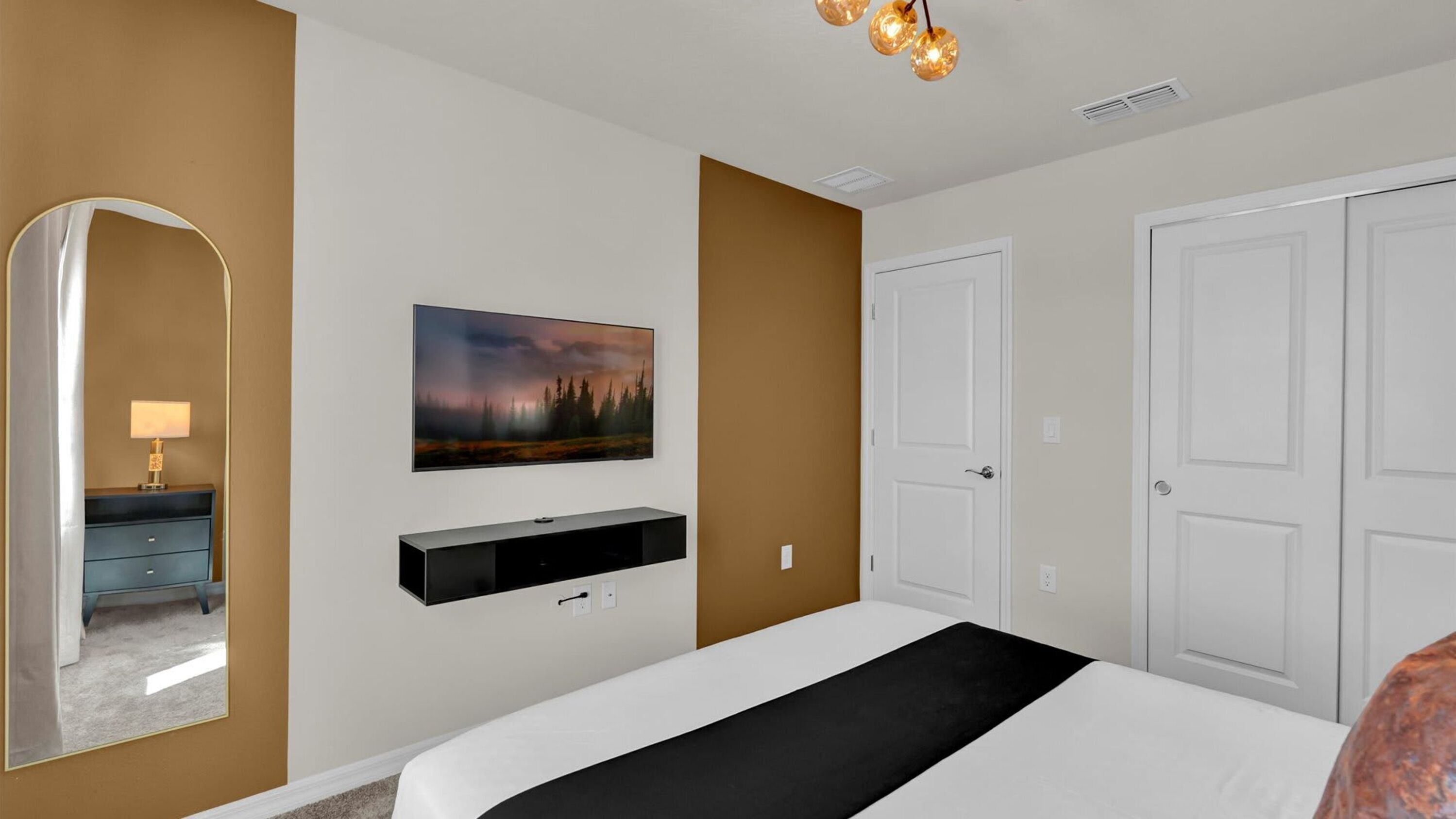 A bedroom with a bed, a bedside table with a lamp, a mounted TV, a painting, and a wardrobe.