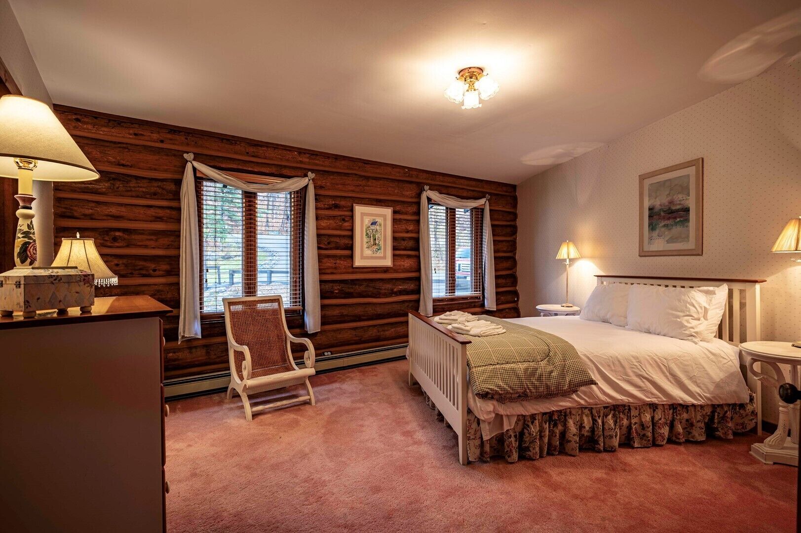 Une chambre confortable avec un mur en bois brut, un lit recouvert d’une courtepointe à motifs floraux, une chaise et deux fenêtres avec des rideaux.