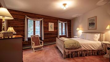 Une chambre confortable avec un mur en bois brut, un lit recouvert d’une courtepointe à motifs floraux, une chaise et deux fenêtres avec des rideaux.