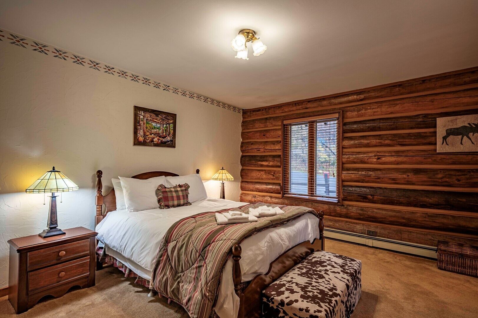 A cozy log cabin bedroom with a wooden bed, two bedside lamps, a framed picture, and a window with blinds.