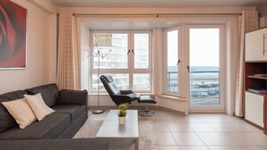 Un salon moderne avec un canapé gris, une table basse, un fauteuil noir et une vue sur la mer grâce à de grandes fenêtres.