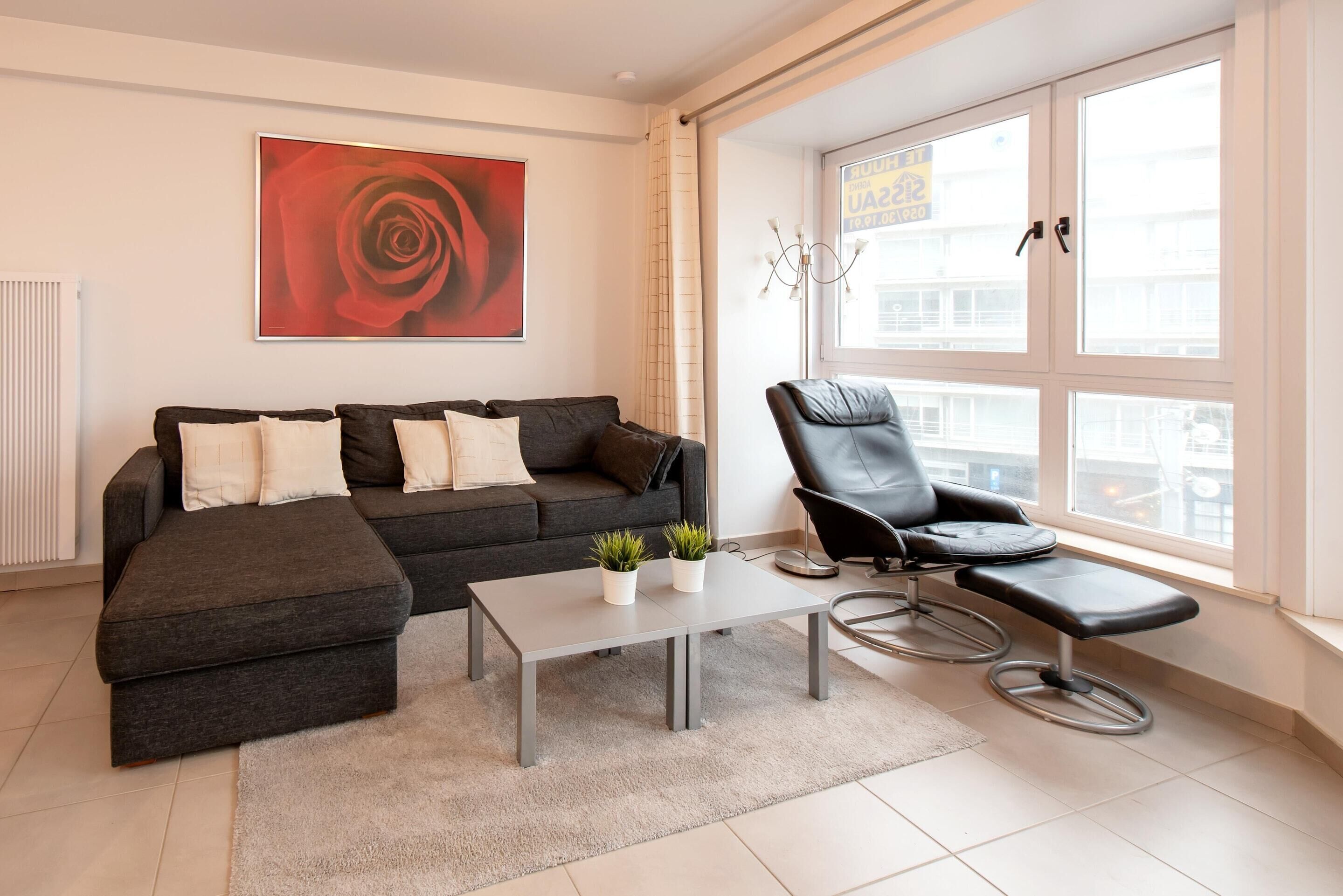A modern living room with a grey sofa, a black leather chair, a small table with plants, and a large window with a view of buildings.