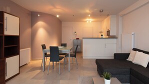 Un salon moderne avec un canapé gris foncé, une table à manger avec des chaises, un coin cuisine doté d’un plan de travail blanc et une petite plante en pot posée sur une table basse.
