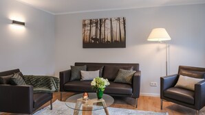 A modern living room with a dark leather sofa, two armchairs, a glass coffee table, and a floor lamp. A painting of trees hangs on the wall. - Garden 5 - Apartment 4 (Braunlage)