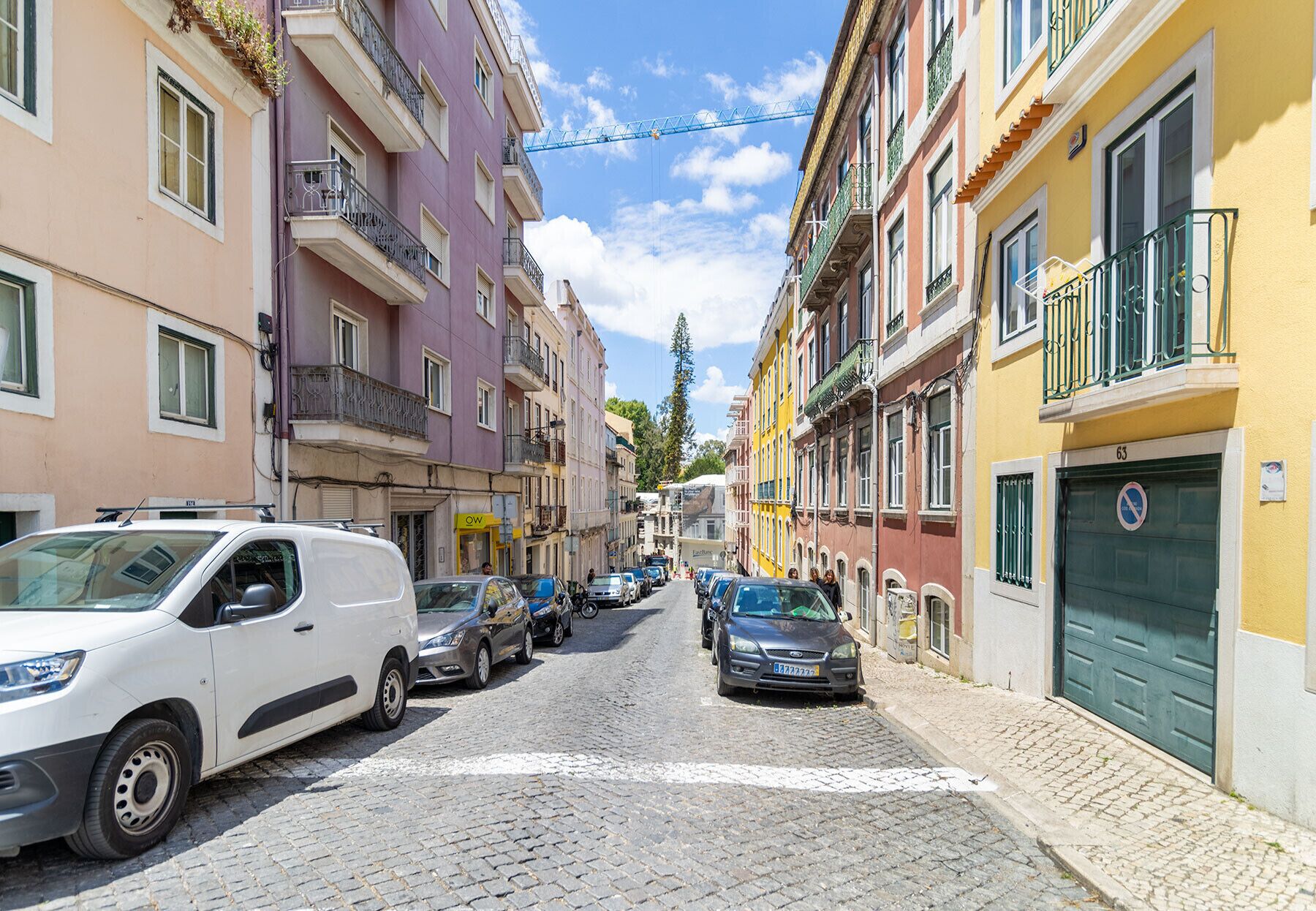 A cobblestone street lined with colorful buildings, parked cars, and a visible 'OW' sign indicating a business.