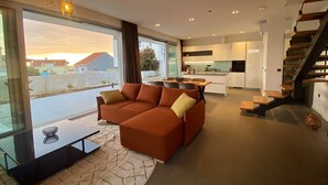 A modern living room with a sofa, a coffee table, and a view of the outside. There is a kitchen area with white cabinets and a staircase.