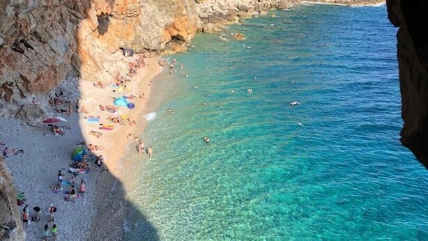 A coastal beach with clear turquoise water, rocky cliffs, and people sunbathing.